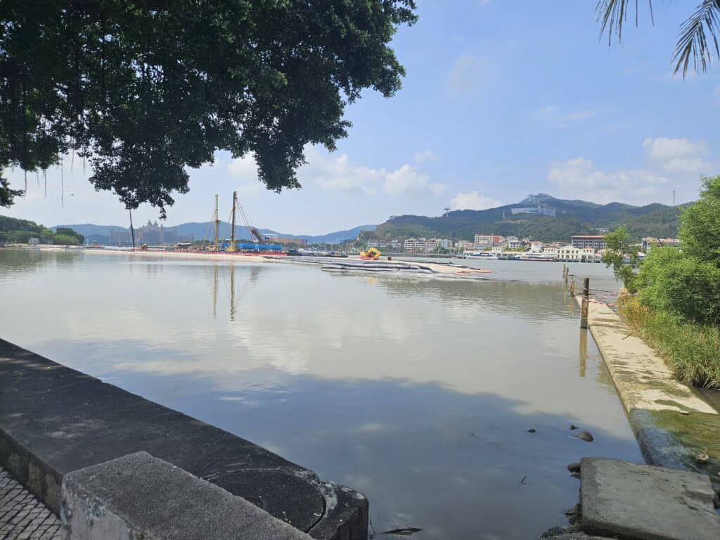 Quiet waterfront scene in Coloane, Macau, with calm water, trees and low-rise buildings backed by green hills.