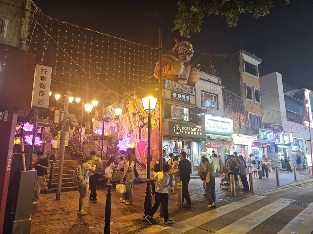 Busy night street in Taipa Village, Macau, with neon lights, restaurants and crowds near the famous staircase mural.
