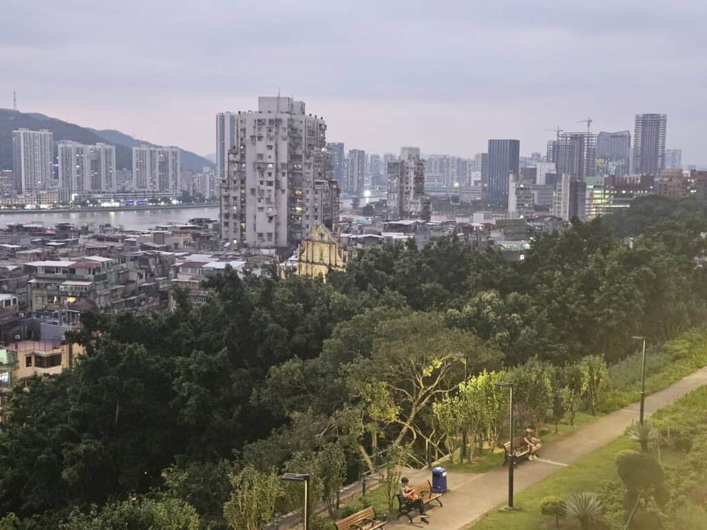 Dusk view over Macau from Monte Forte, with the illuminated Ruins of St Paul’s visible among apartment buildings and trees.