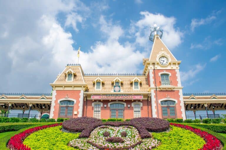 Hong Kong Disneyland entrance with its red brick clock tower and Mickey Mouse flower display under a bright blue sky.